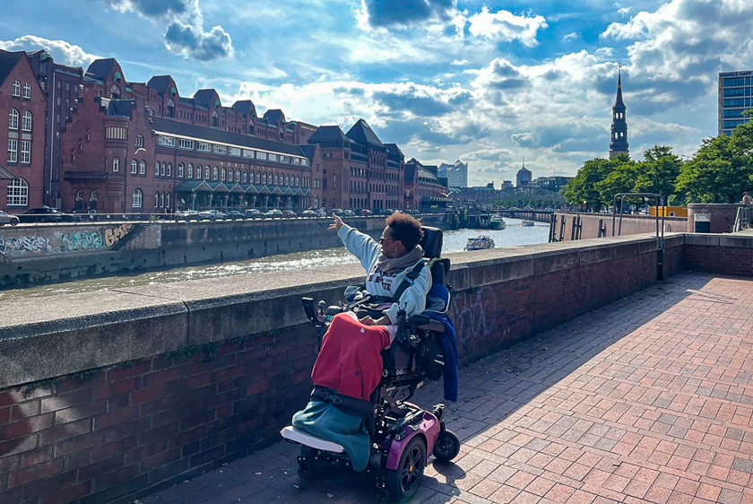 Eine Dame im Elektrorollstuhl steht an einem Fluss in einer Innenstadt und zeigt auf ein Gebäude auf der anderen Seite des Flusses.