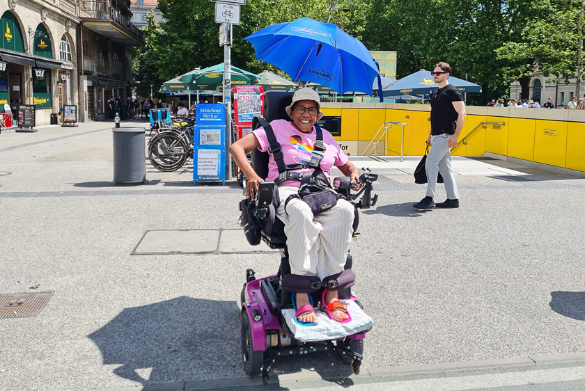 Eine strahlend lachende Dame sitzt in ihrem pinken PR30/II Elektrorollstuhl, leicht hochgefahren, in einer Stadt. An ihrem Rollstuhl ist ein geöffneter Sonnenschirm befestigt worden. 