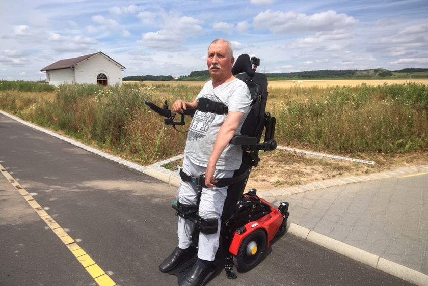 Ein älterer Herr steht in seinem PR40 Stehrollstuhl auf einem Weg in der Natur. Um ihn herum bei Sonnenschein nur Wiesen und Felder, sowie links ein kleines Häuschen.