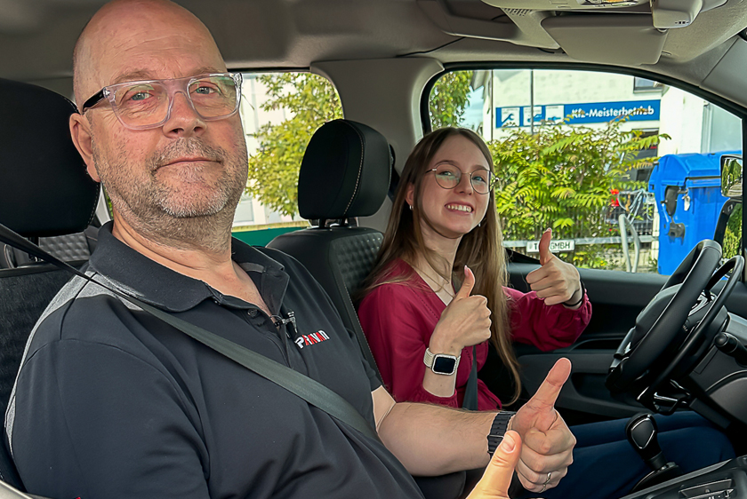 In einem Auto sitzt eine junge Frau am Steuer und streckt mit einem großen lächeln beide Daumen in die Luft. Auf dem Beifahrersitz, sitzt ein Fahrlehrer der Fahrschule Paravan und streckt ebenfalls mit einem lächeln einen Daumen in die Luft.