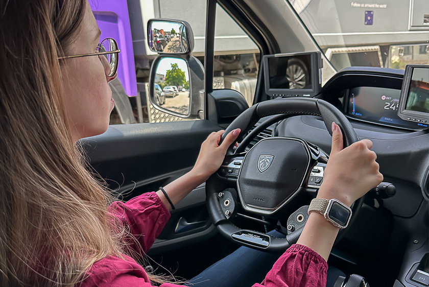 Über die rechte Schulter schaut man einer jungen Dame dabei zu wie sie Autofährt. Beide Hände befinden sich am Lenkrad.