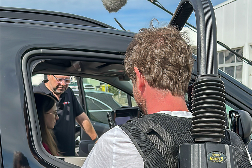 Ein Kameramann filmt durch ein geöffnetes Beifahrerfenster eines Autos. Er filmt eine Junge Dame, die auf dem Fahrersitz sitzt und neben ihr an der Tür steht ein Mann und zeigt ihr etwas.