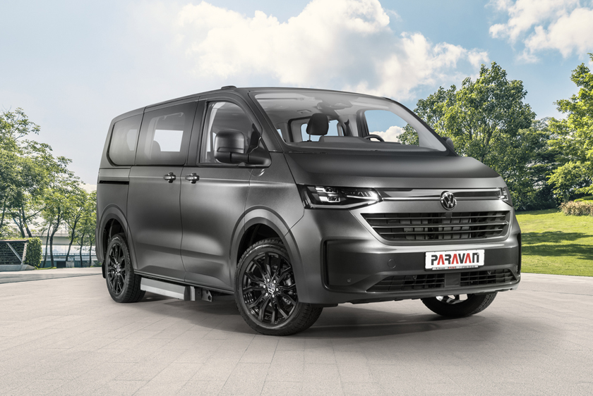 Die VW New Caravelle stht auf dem Hof des PARAVAN Mobilitätspark, fotografiert bei Sonnenschein von vorne rechts. Das Auto ist in einem dunklen grau und hat ein PARAVAN Logo vorne im Kennzeichenhalter.