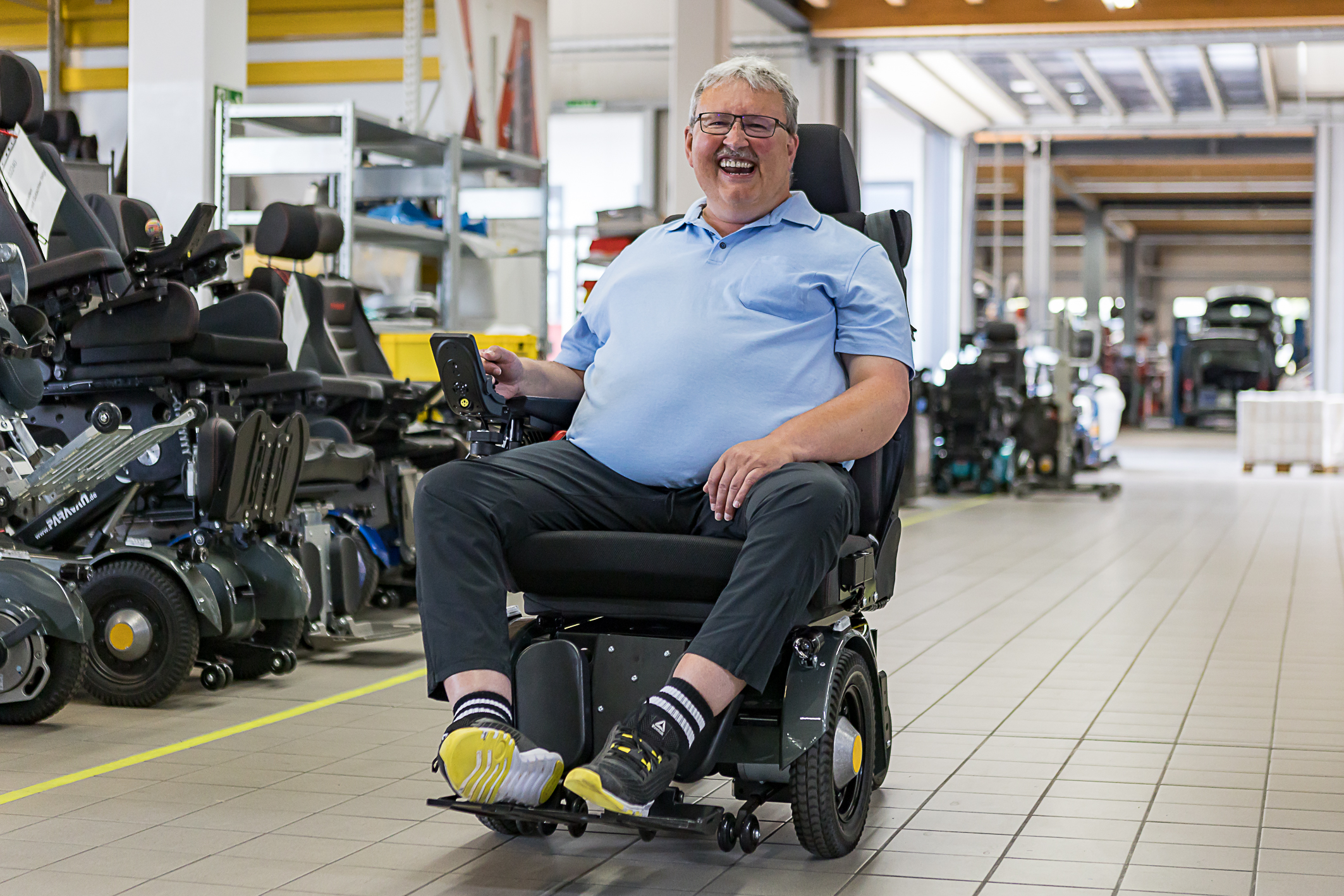 Ein älterer Mann sitz in seinem Elektrorollstuhl und fährt auf einem Gang in einer Halle. Links stehen noch weitere Rollstühle. Der Mann lacht, strahlt und ist sehr glücklich.
