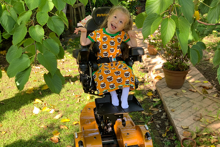 Ein Junges Mädchen sitzt im Garten unter einem Baum, mit orangenen Kleid passend zu ihrem Orangenen Elektrorollstuhl von Paravan. Sie hat den Kopf leicht nach rechts geneigt die Hände in einer Pose und streckt frech die Zunge raus. 