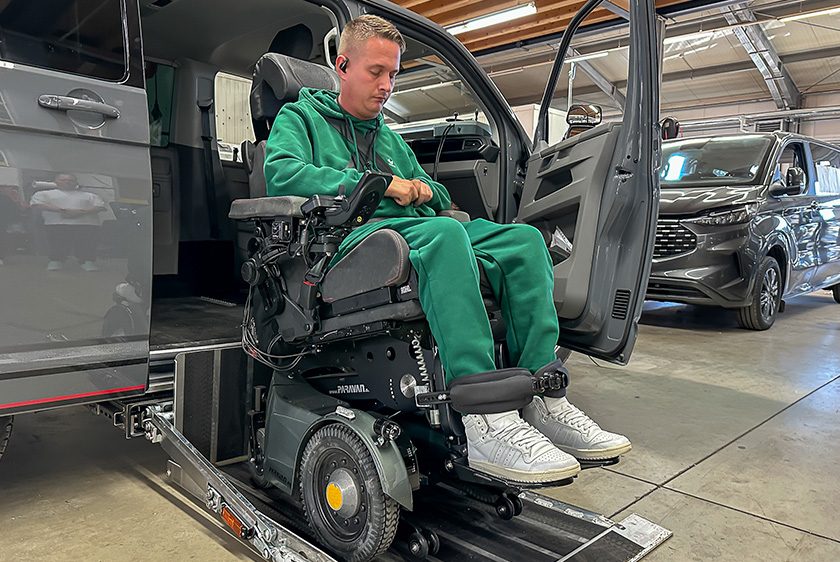 Ein Mann im grünen Sportklamotten sitzt in seinem Elektrorollstuhl. Beide zusammen stehen auf dem Kassettenlift von Paravan an einem grauen Auto. 