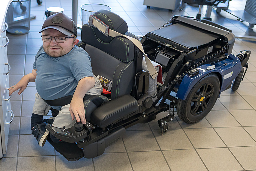 Ein Kleinwüchsiger Mann sitzt in seinem dunkelblauen PR25 Rollstuhl, der runter gefahren wurde bis zum Boden, damit er an die untere Schublade des Schreibtisches kommt.
