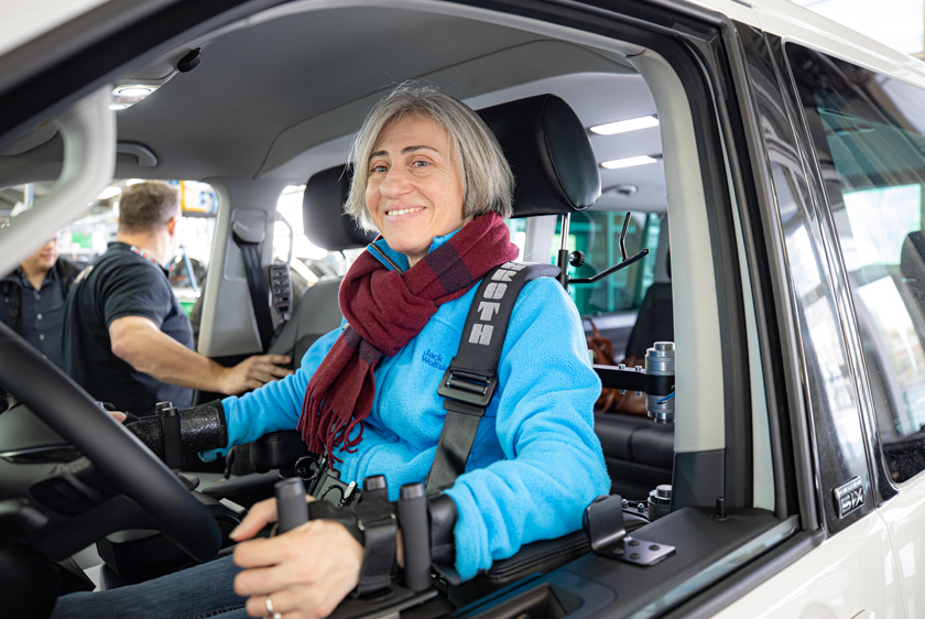 Eine etwas ältere Dame mit grauen haaren einem dunkelroten Schal und blauer Jacke sitzt am Steuer eines Fahrzeugs. Sie hält in ihrer linken Hand ein Dreizack Joystick und lächelt. Im Hintergrund reden zwei Personen miteinander. 