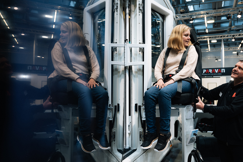 Eine Dame sitzt in einem Lift welcher hoch gefahren wurde um leicht in ein Wohnmobil zu kommen. Von rechts hilft ihr noch ein Mitarbeiter der Paravan GmbH. Links spiegelt sich die Dame im Wohnmobil.