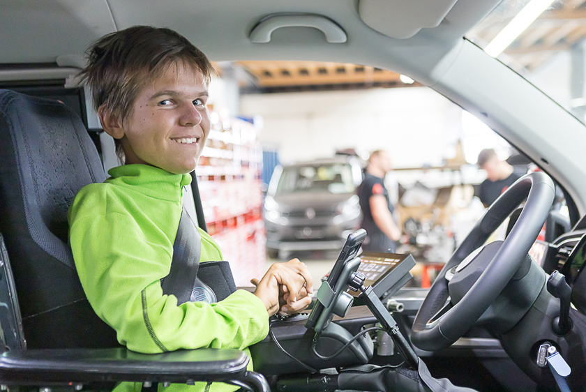 Ein junger Mann sitzt am Steuer eines PKWs. Mit seinem grünen Pullover und einem lächeln schaut er zur Beifahrerseite. Vor seinem Lenkrad befinden sich ein Touch Display und eine Handyhalterung.