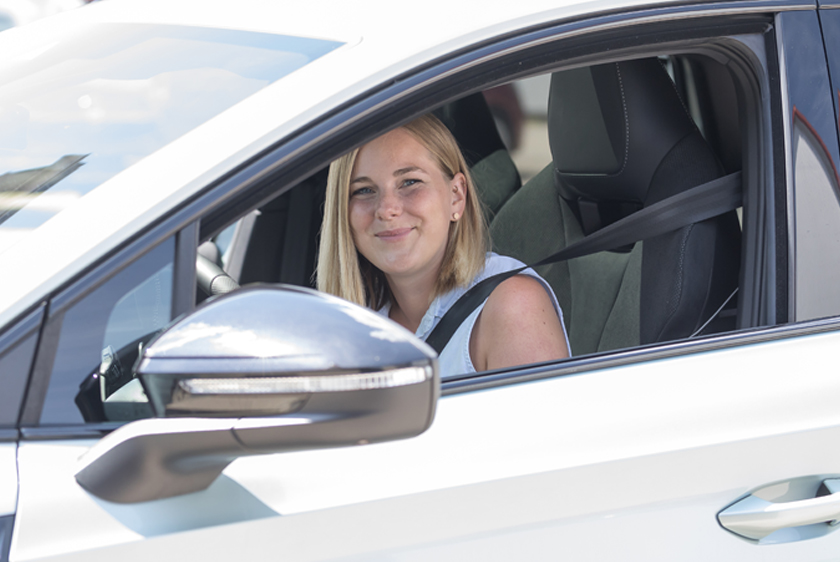 Eine nett lächelnde junge Frau mit blonden glatten Haaren schaut zum geöffneten Fahrerfenster hinaus.
