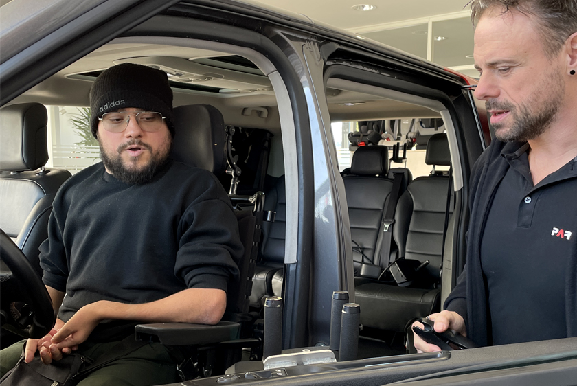 Ein Kunde sitzt in einem Auto am Fahrerplatz, rechts bei geöffneter Türe steht ein Mitarbeiter von Paravan und hält einen Joystick fest.