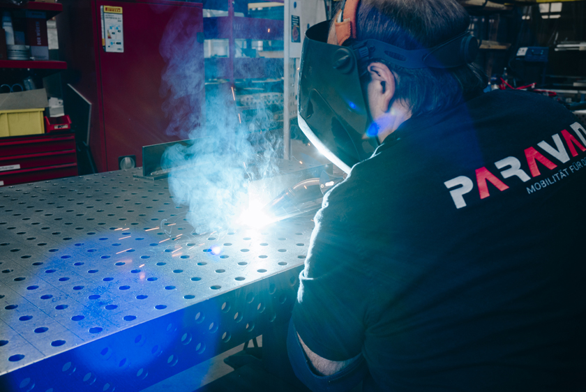 PARAVAN Mitarbeiter beim Schweißen in der Werkstatt, aufgenommen von hinten mit sichtbarem PARAVAN Logo auf dem Shirt.