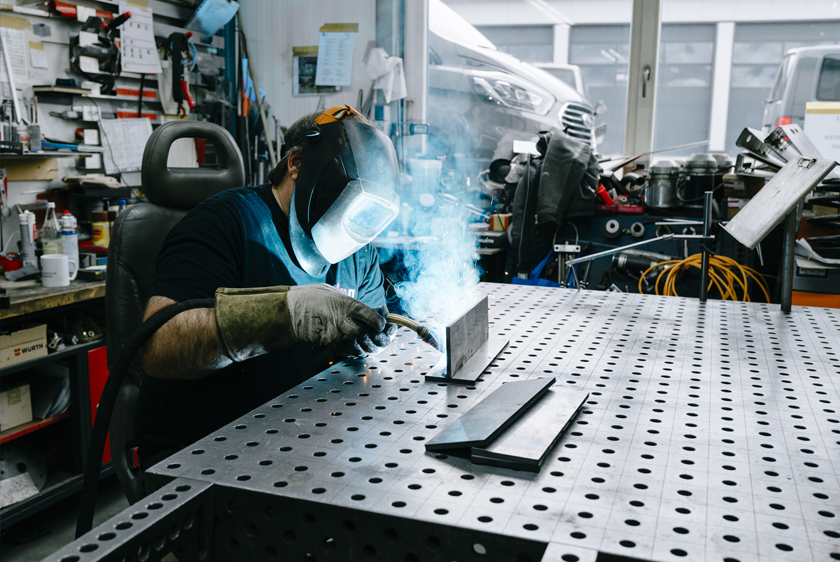 PARAVAN Mitarbeiter mit Schweißhelm beim Schweißen eines Metallbauteils an einem Arbeitstisch.