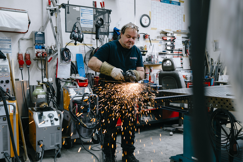PARAVAN Mitarbeiter beim Schleifen eines Metallbauteils in der Werkstatt, Funken sprühen in Richtung Boden.